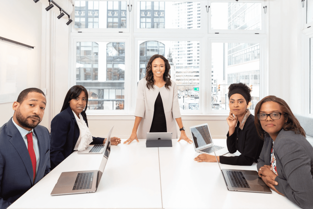 Boardroom meeting with a woman standing in the center of the photo, flanked on either side by a total of four people.