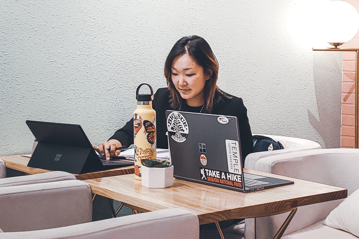 Woman sitting at a corner table with a black Microsoft laptop in front of her. You can also see a water bottle, wall decorations, and a trendy Windows laptop next to her. Apprenticeships Microsoft