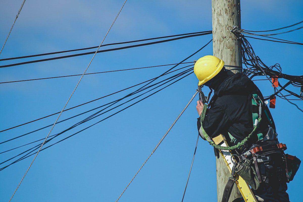 A man wearing a yellow helmet and tool belt standing at the top of a telephone pole, fixing power lines How to Become an Electrical Engineer