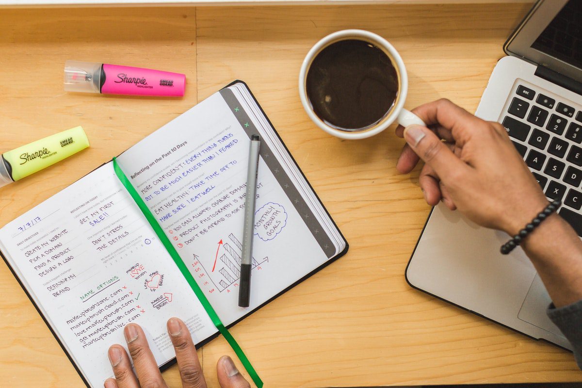  A person holding a cup of coffee and reading from a notebook next to a laptop how to become a content marketer