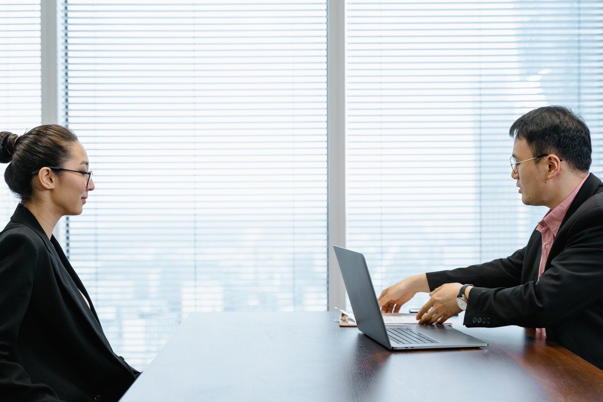 A man at a desk with a laptop interviewing a woman wearing glasses.