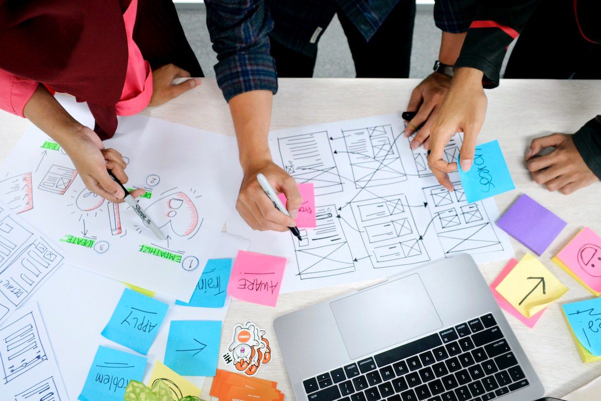 Three people are gathered around a table filled with design plans and notes. UX Designer Interview Questions and Answers