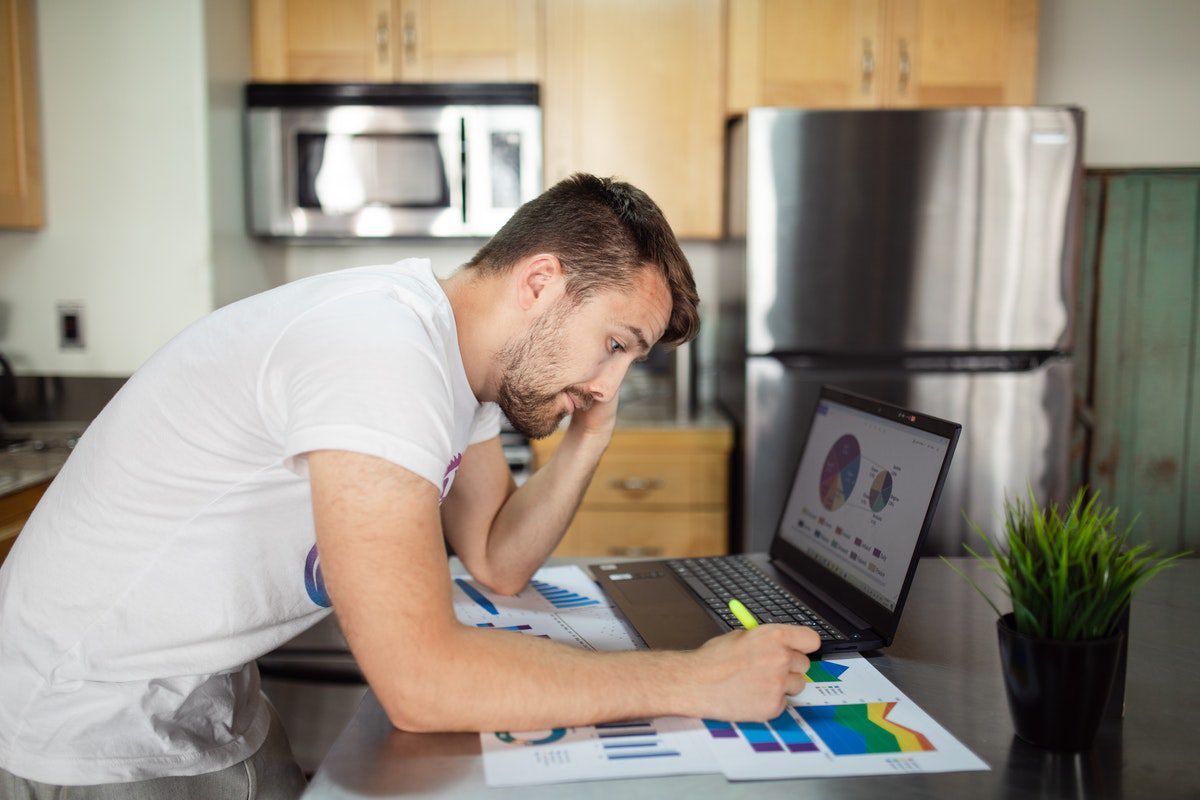 Man working at a kitchen counter on a laptop and paperwork showing several colorful graphs. Supply Chain Management Master's Degrees