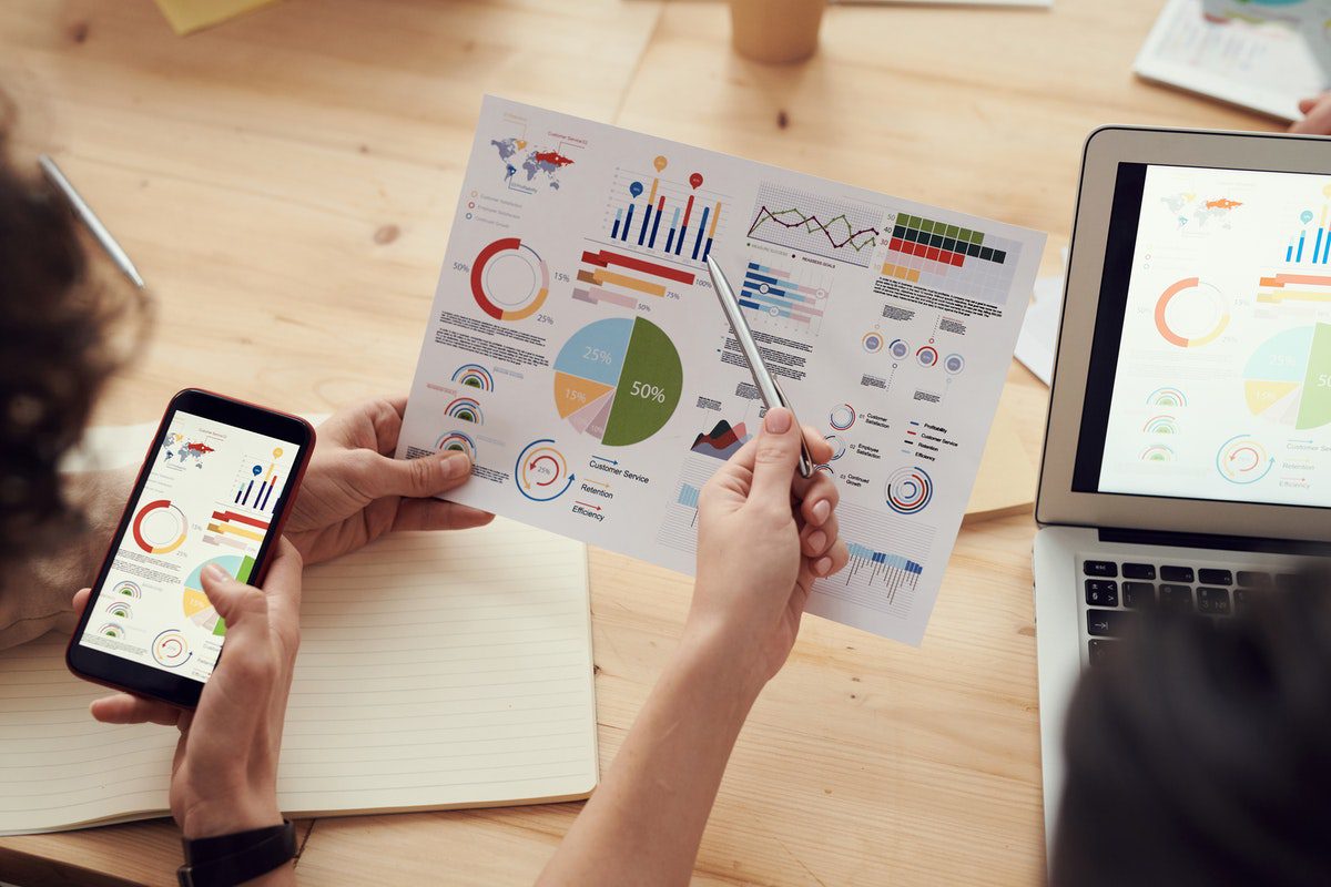 People looking at charts on paper, a phone, and a laptop. Excel Projects