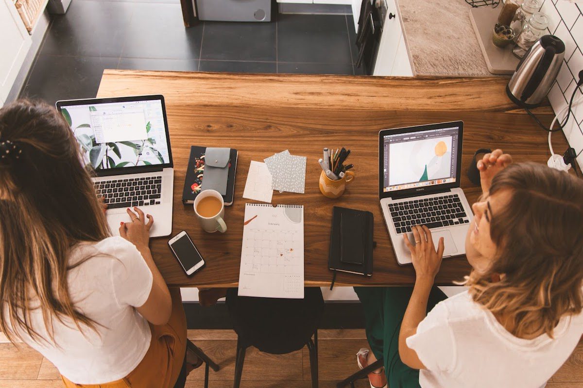 Two ladies working on their laptops and chatting.