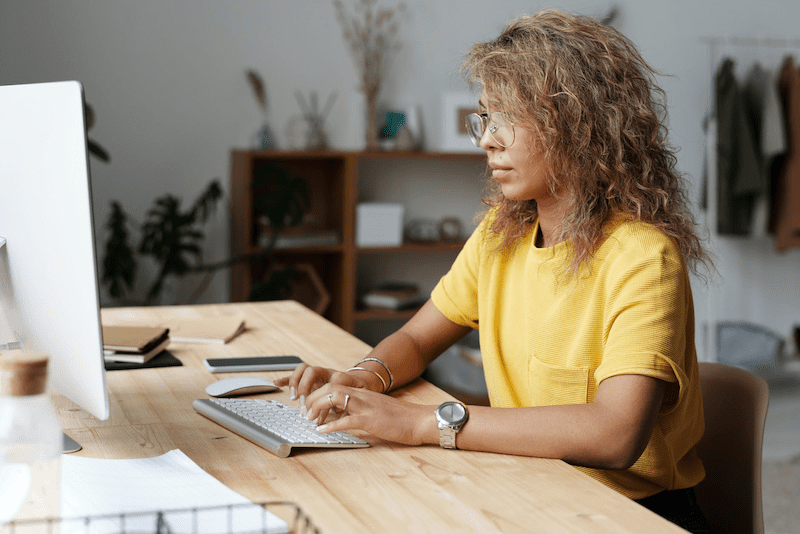 A woman working on a C# project at a wooden desk with a monitor and wireless keyboard.