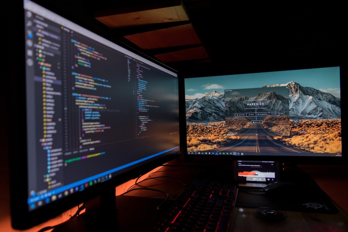  Two black computer monitors on a black table with a phone, white keyboard, a mouse, and a pair of black headphones on it. Python for Business