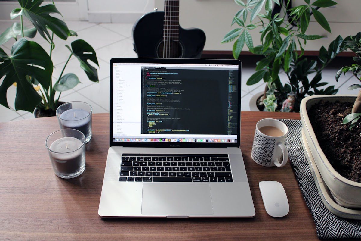 A laptop displaying JavaScript code in a text editor on its screen alongside with unlit candles, a mug with coffee, on a desk surrounded by house plants.