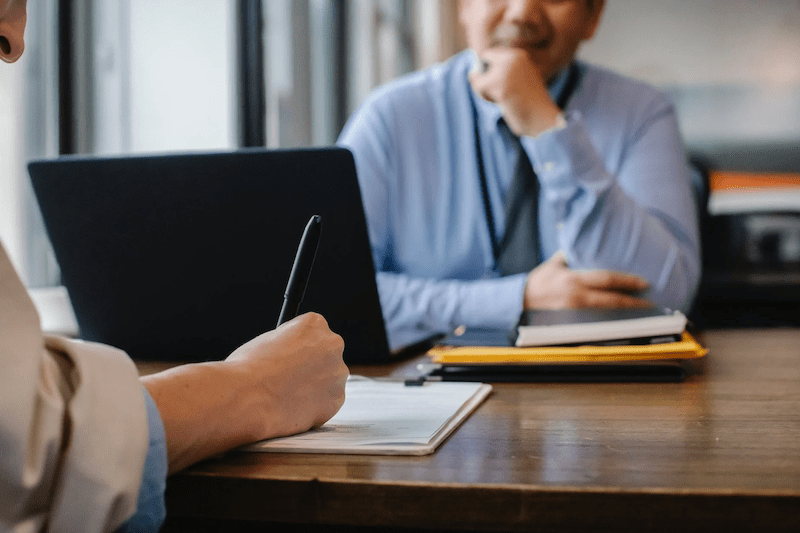 A man in front of a laptop sitting across from a woman who is writing on a paper.