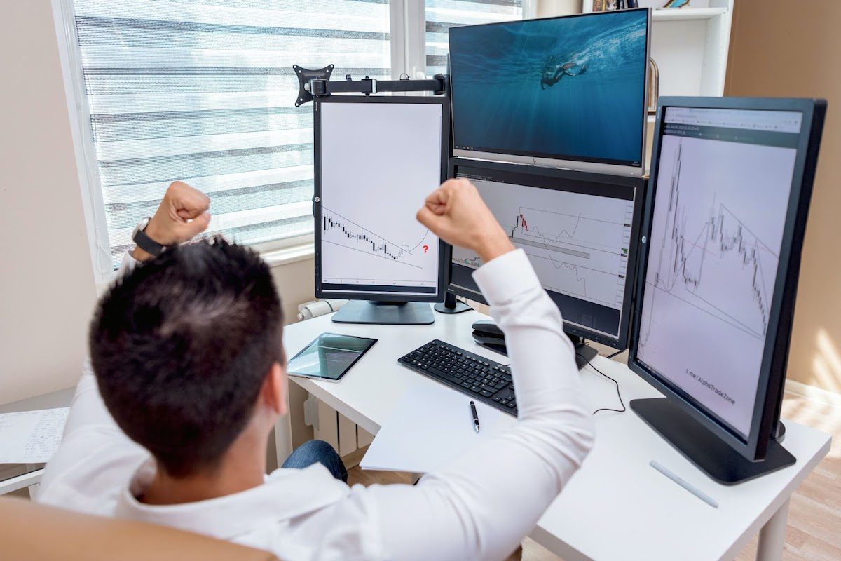 A man sitting in front of four computer screens with his hands in jubilation