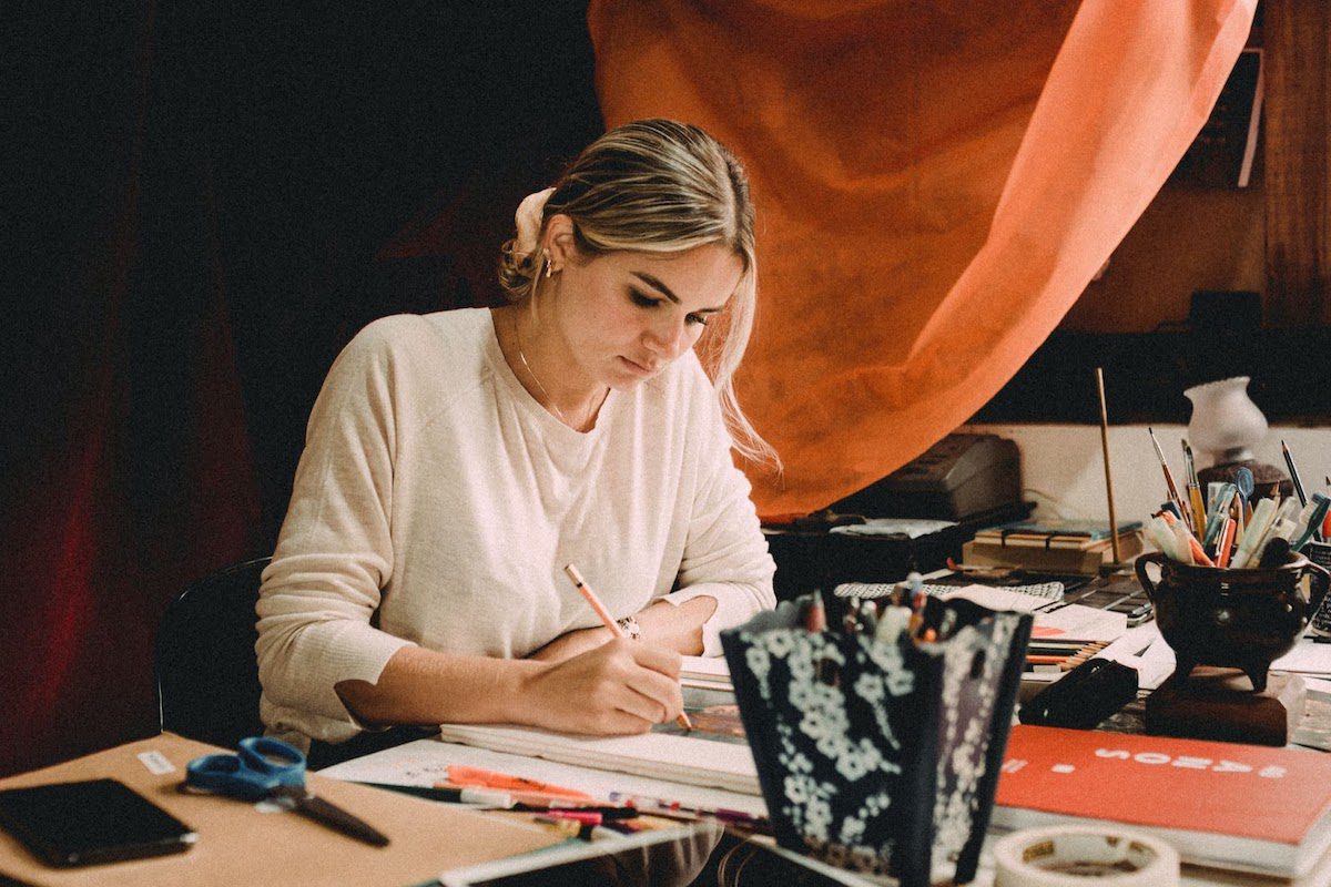 Woman sitting on a desk full of art materials working on a piece.