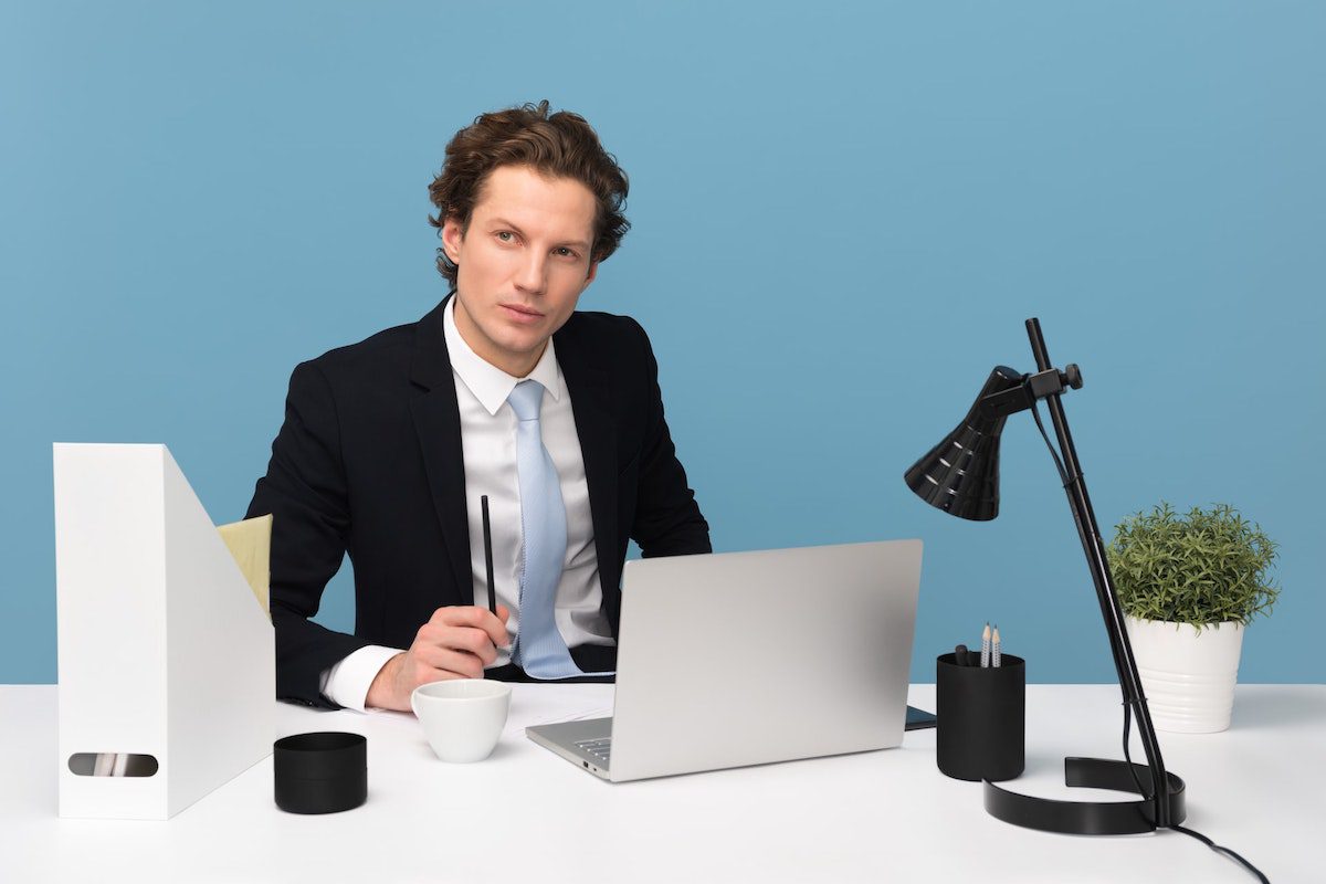 A man wearing a coat and tie sitting on his desk with a laptop, lamp, and a plant. Accounts Manager Cover Letter