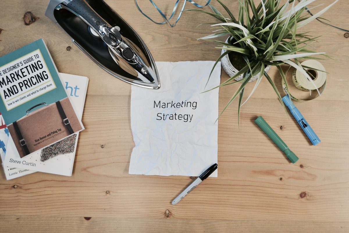 A table with a white paper with “marketing strategy” written on it placed next to a textbook, an iron, pens, and a white plant pot. Marketing Manager Job Satisfaction