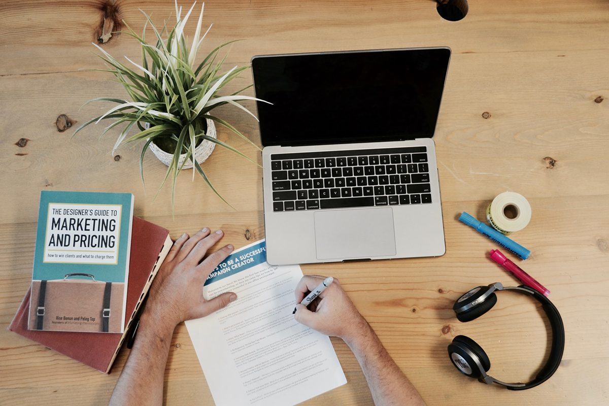 A person writing on a sheet, next to a laptop, books, headphones, and a plant on a table. Marketing Coordinator Cover Letter