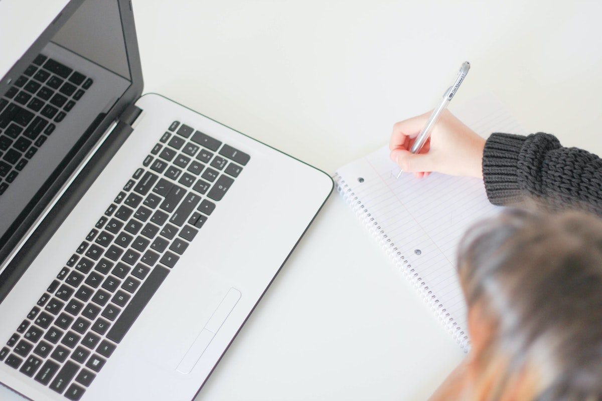 A woman writing in her notepad next to a laptop. Operations Analyst Cover Letter