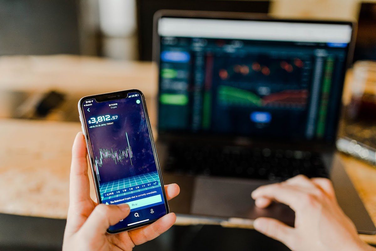 Someone holding their smartphone displaying financial graphs while working on their computer