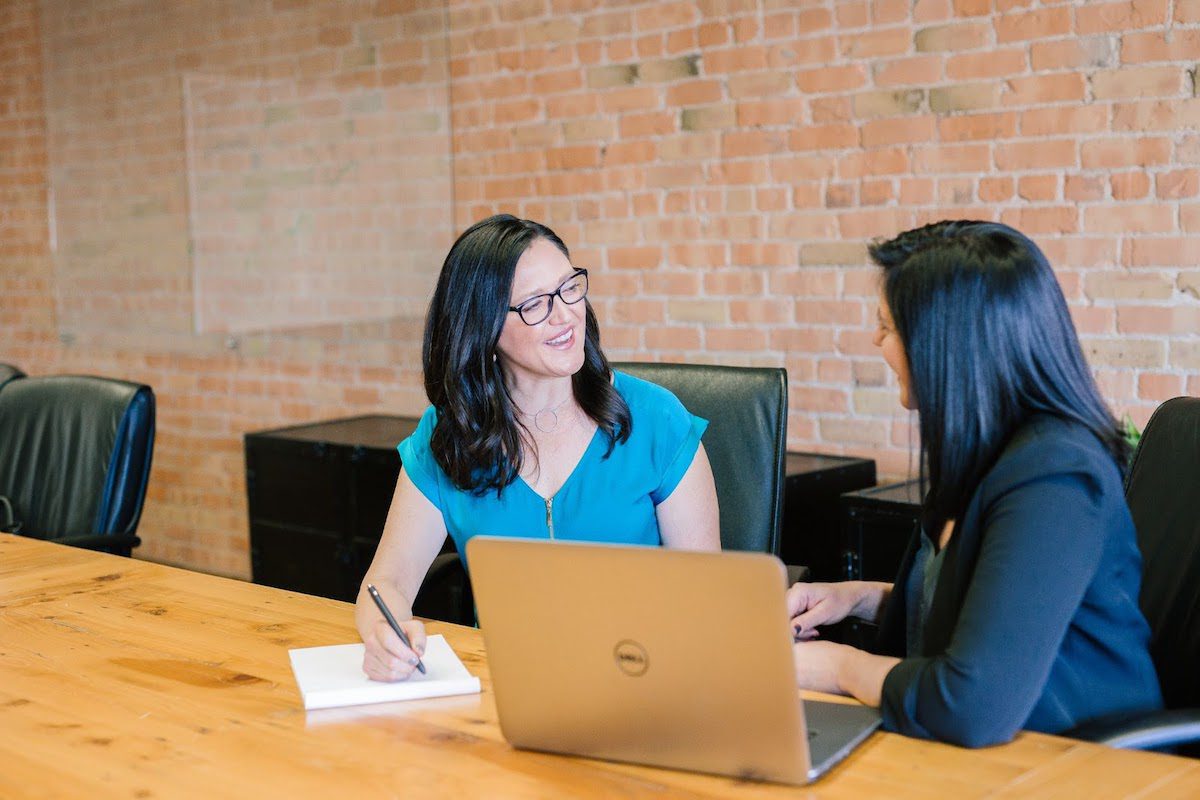 A picture of a woman in a teal shirt taking notes from a woman in a suit.