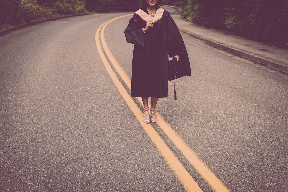 A happy woman wearing graduation regalia and standing on a road