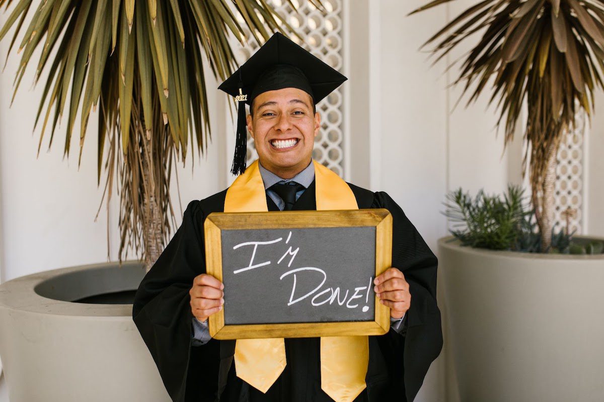 Man in a graduation cap and gown holding a small blackboard with ‘I’m done!’ written on it in chalk.