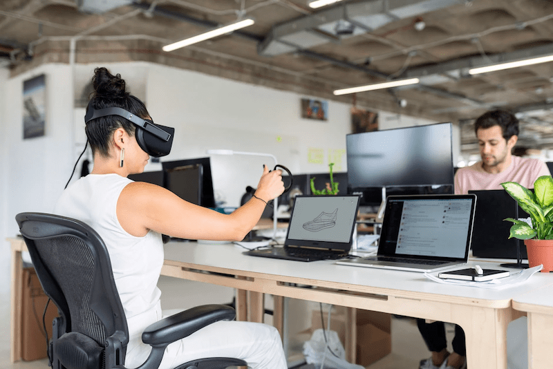 A woman wearing a VR headset is seated in front of a laptop that has a shoe design on the screen.