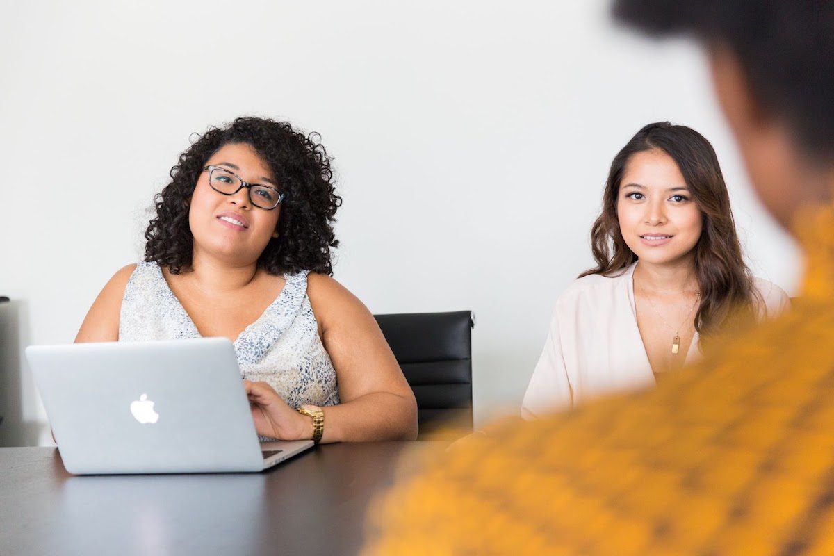 Three people in a meeting sitting at a desk, one of which has a laptop