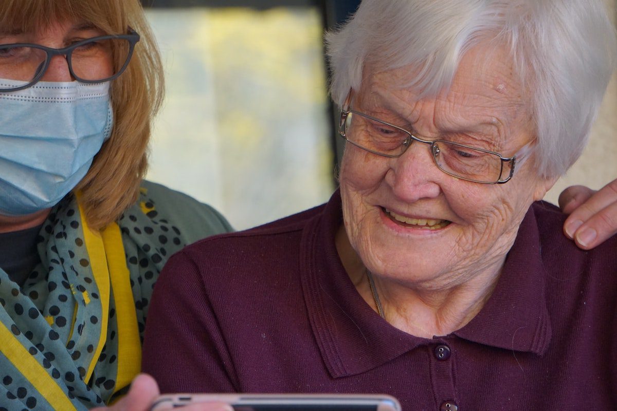 A person in a purple polo shirt wearing glasses next to another person wearing a mask showing them their phone.  How Much Can You Make on Care.com?