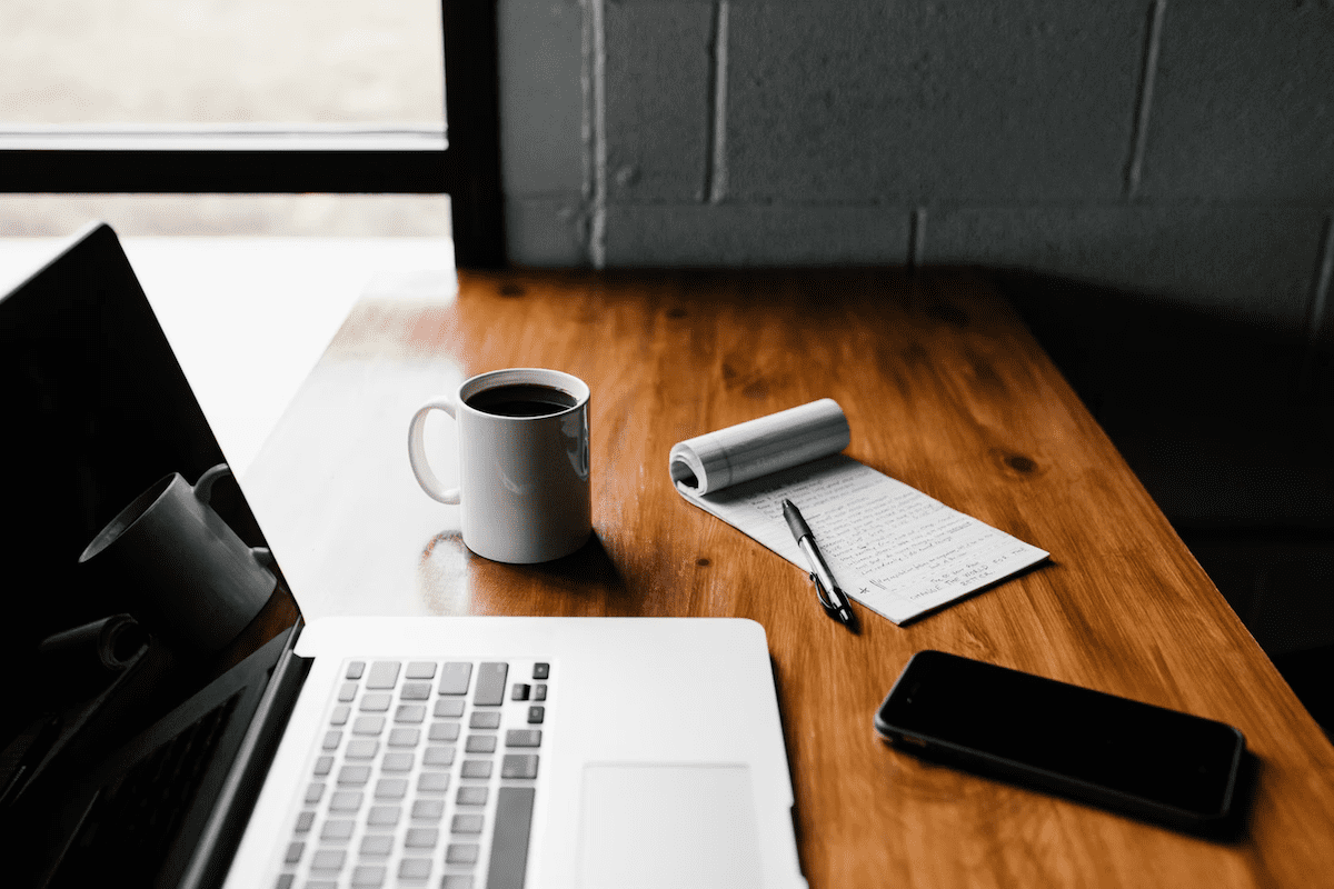 A photo of a laptop on a table alongside a phone, paper, pen, and coffee used for their full-time job. 