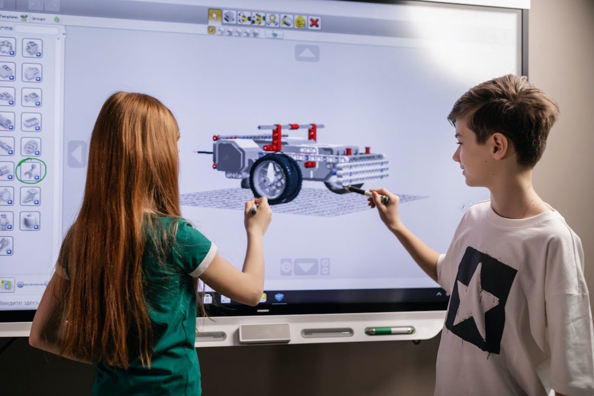 A boy and a girl standing in front of a screen showing an image of a robot.