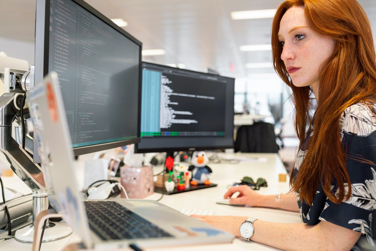 A woman sitting at a desk looking at a laptop with two monitors in front of her