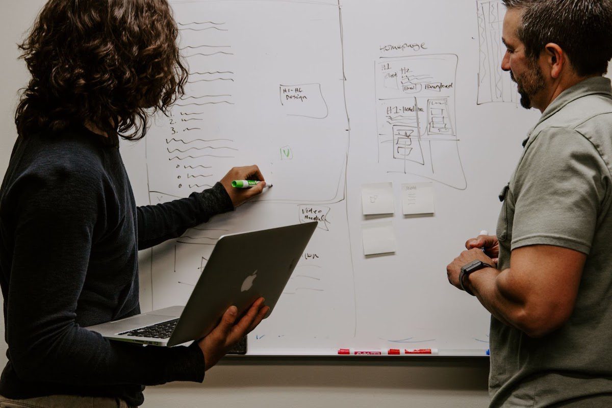 A man in a grey shirt making game development plans on a whiteboard with a woman in a black shirt