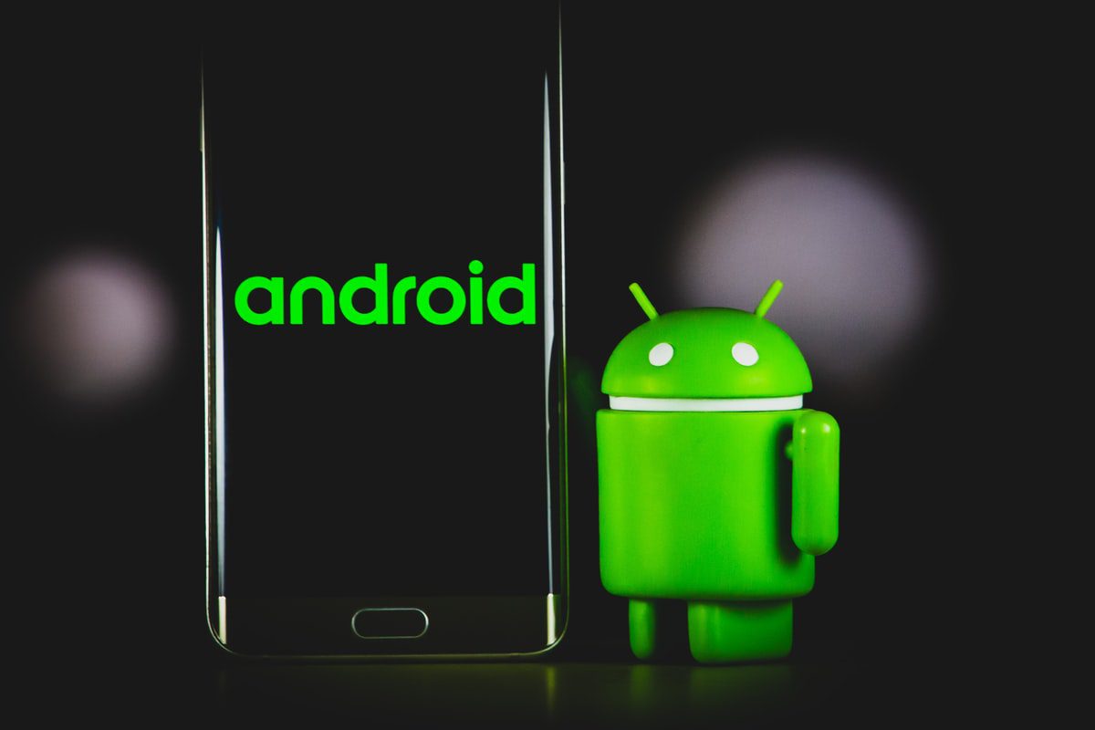A picture of a green Android mascot next to a black phone with Android written on it on a black background. 