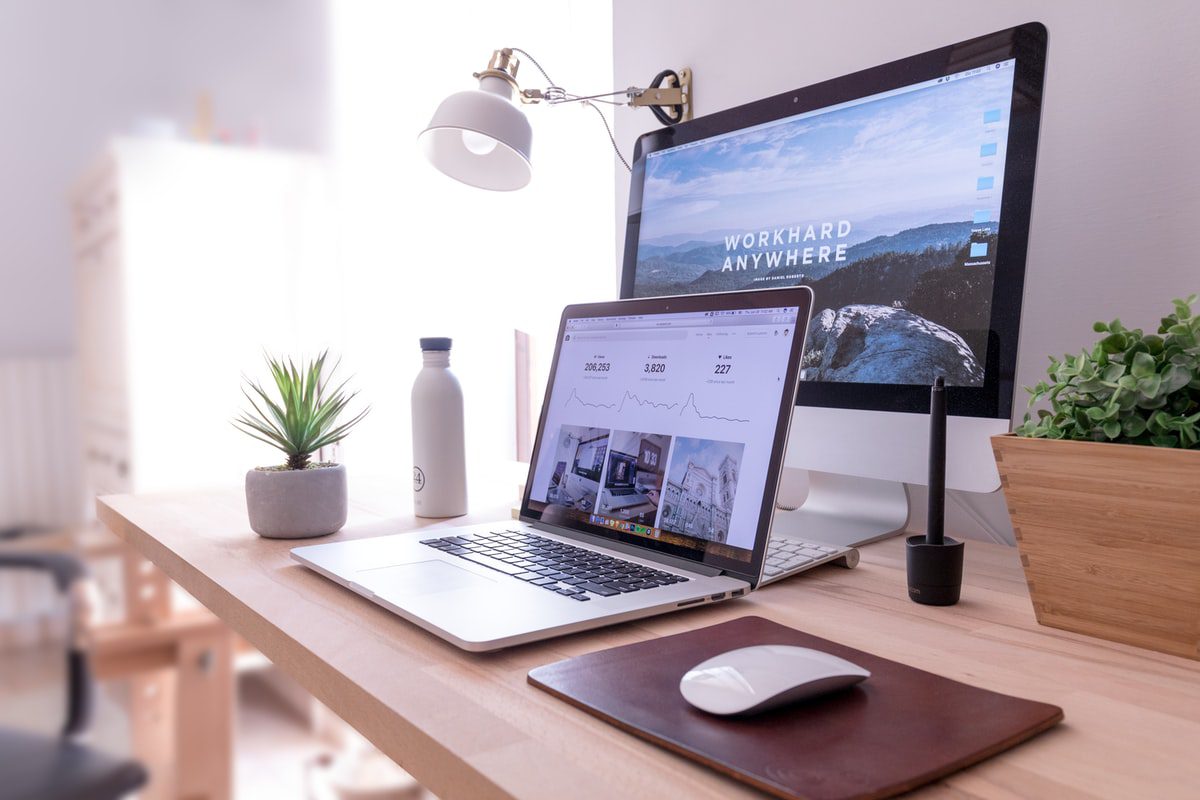 A MacBook Pro, iMac, and Magic Mouse on a table next to two plants, a lamp, and a water bottle. 