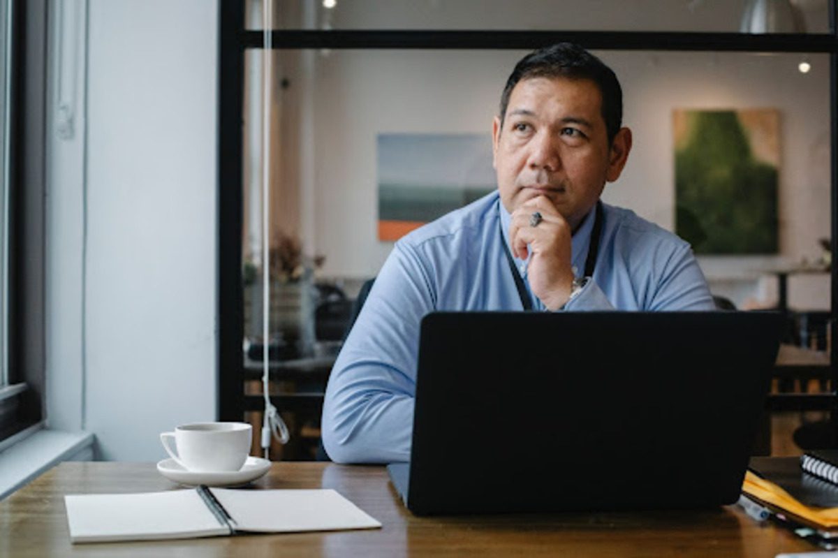 A middle-aged person working on a black laptop with a large cup of coffee and an open notebook.