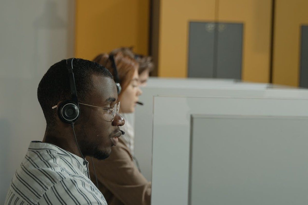 A row of people sitting at desks looking toward a computer screen.