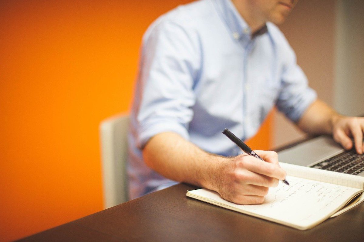 A man writing in a notebook while scrolling on a laptop. Java Remote Jobs