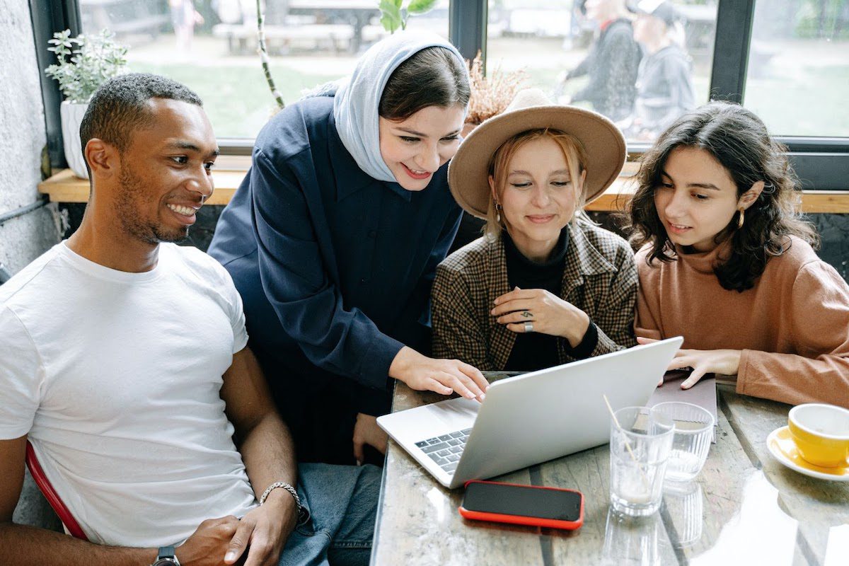 Three women and one man looking at a laptop screen and smiling while sitting at a cafe table.