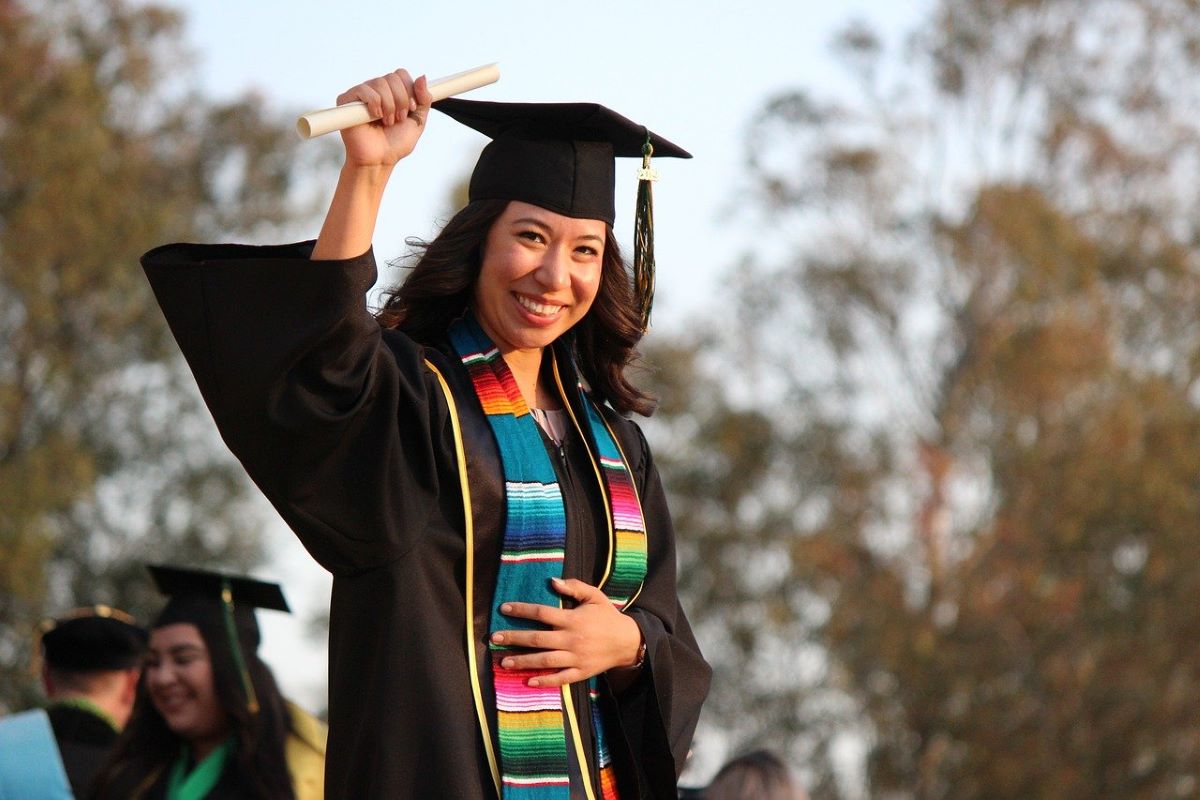 A college student wearing a graduation robe, graduation cap, and holding a degree scroll waving at her graduation ceremony