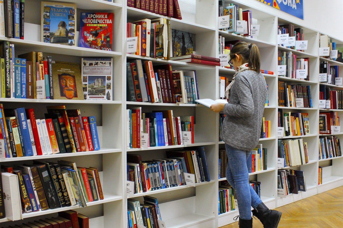 A female student standing beside a bookshelf in a library reading a book.