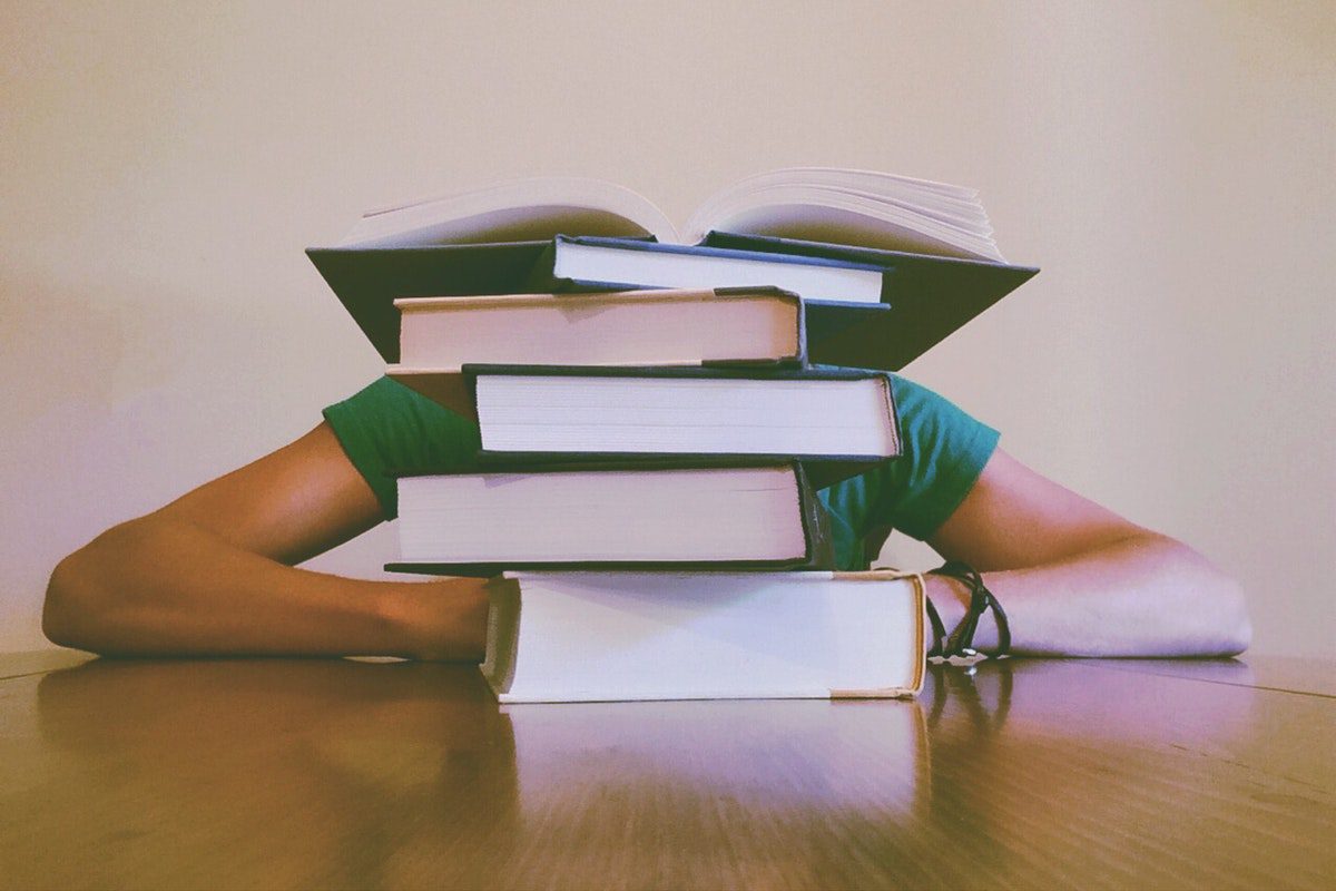 Student at a table with a stack of books covering their head. Accredited Online Colleges In Arkansas