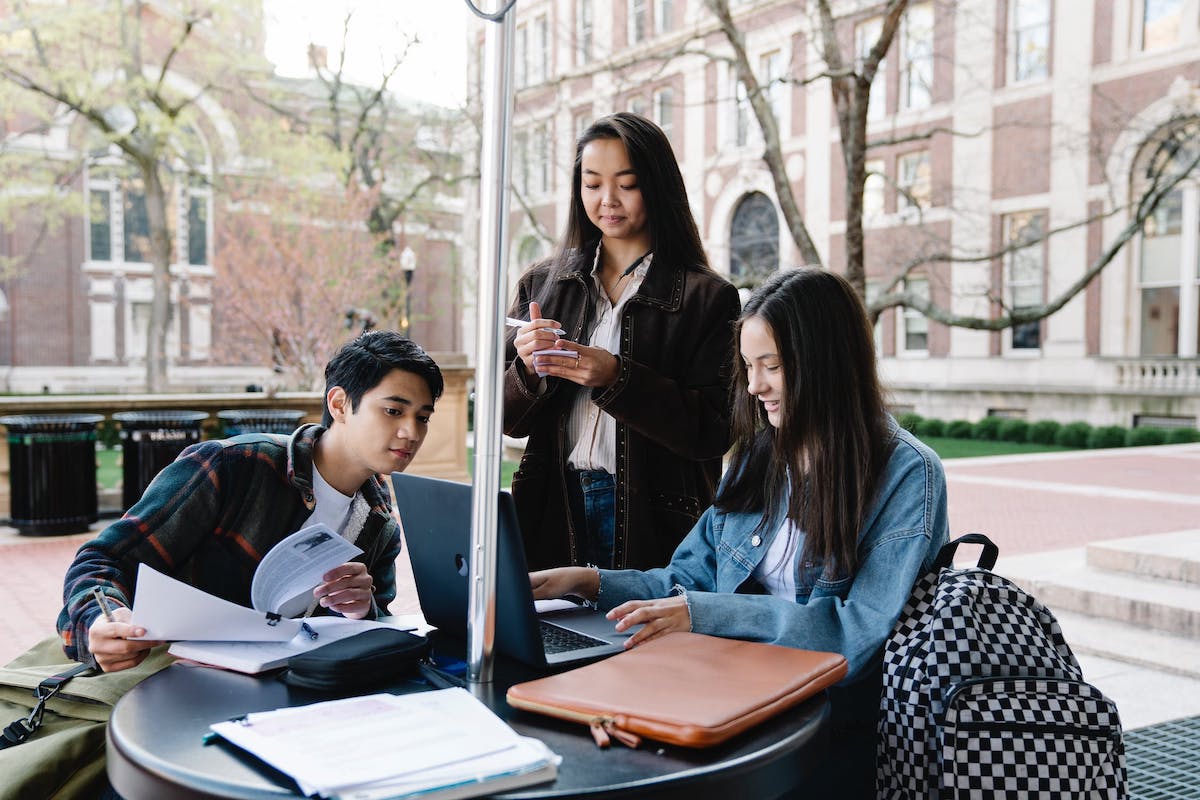 Three undergraduate students study while looking at a laptop in an outdoor college environment. Accredited Online Colleges In Michigan