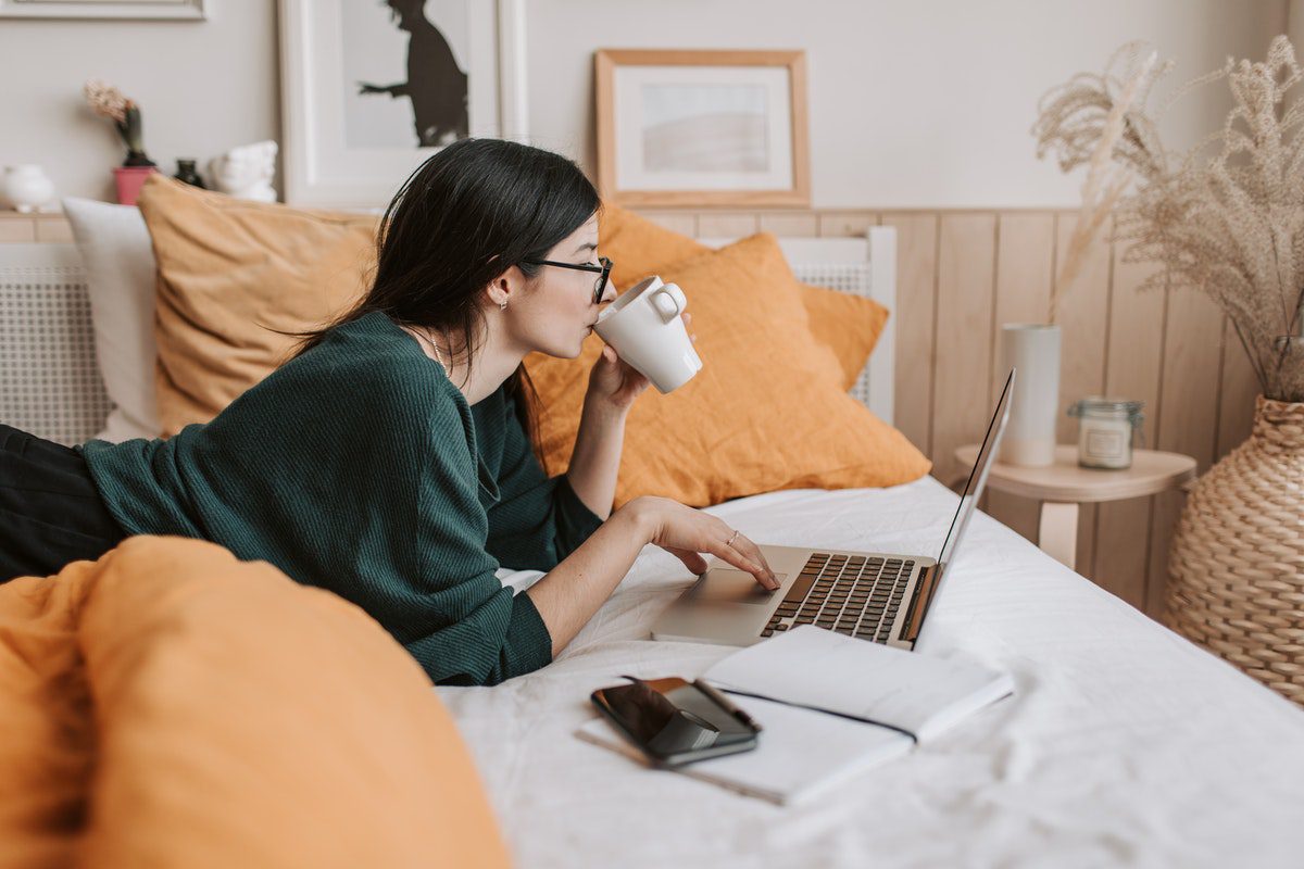 Student lounging on her bed while scrolling through her laptop and drinking from a mug. Accredited Online Colleges In New York