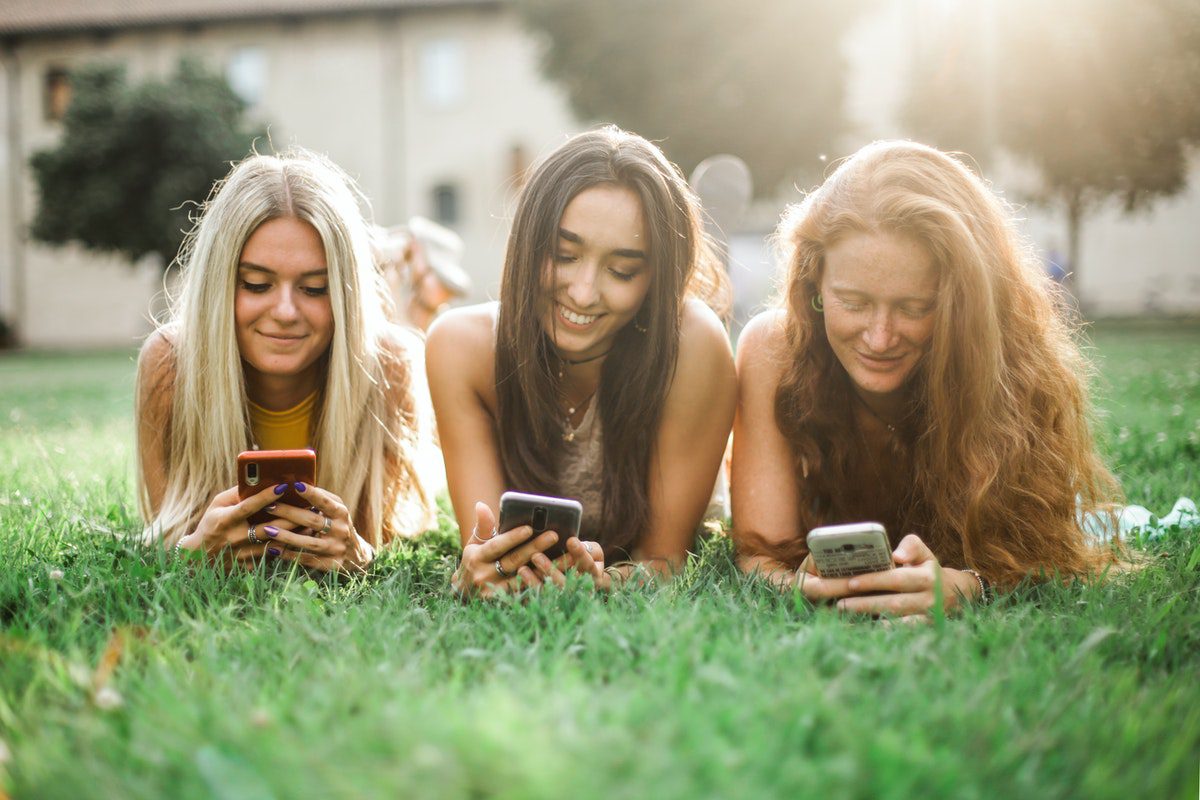 Three girls laying on a lawn smiling using their smartphones. Trends In Social Media
