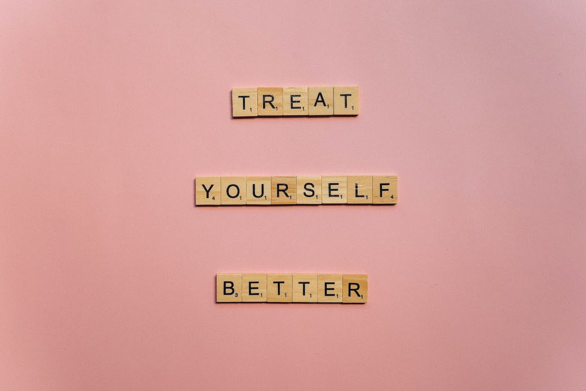 Brown wooden scrabble tiles spelling out “Treat Yourself Better” on a pink background.