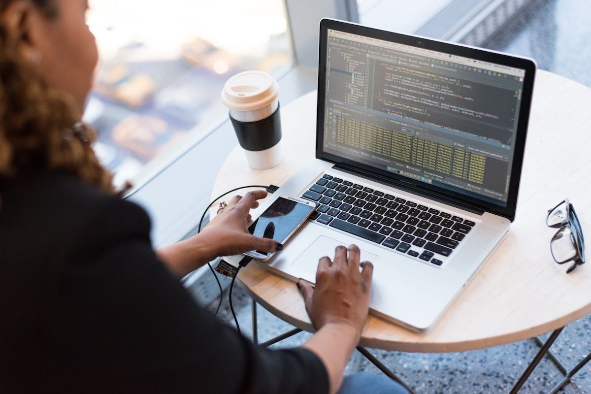 A mobile developer analyzing lines of code on a laptop on a table with a disposable coffee cup, a smartphone, and glasses.