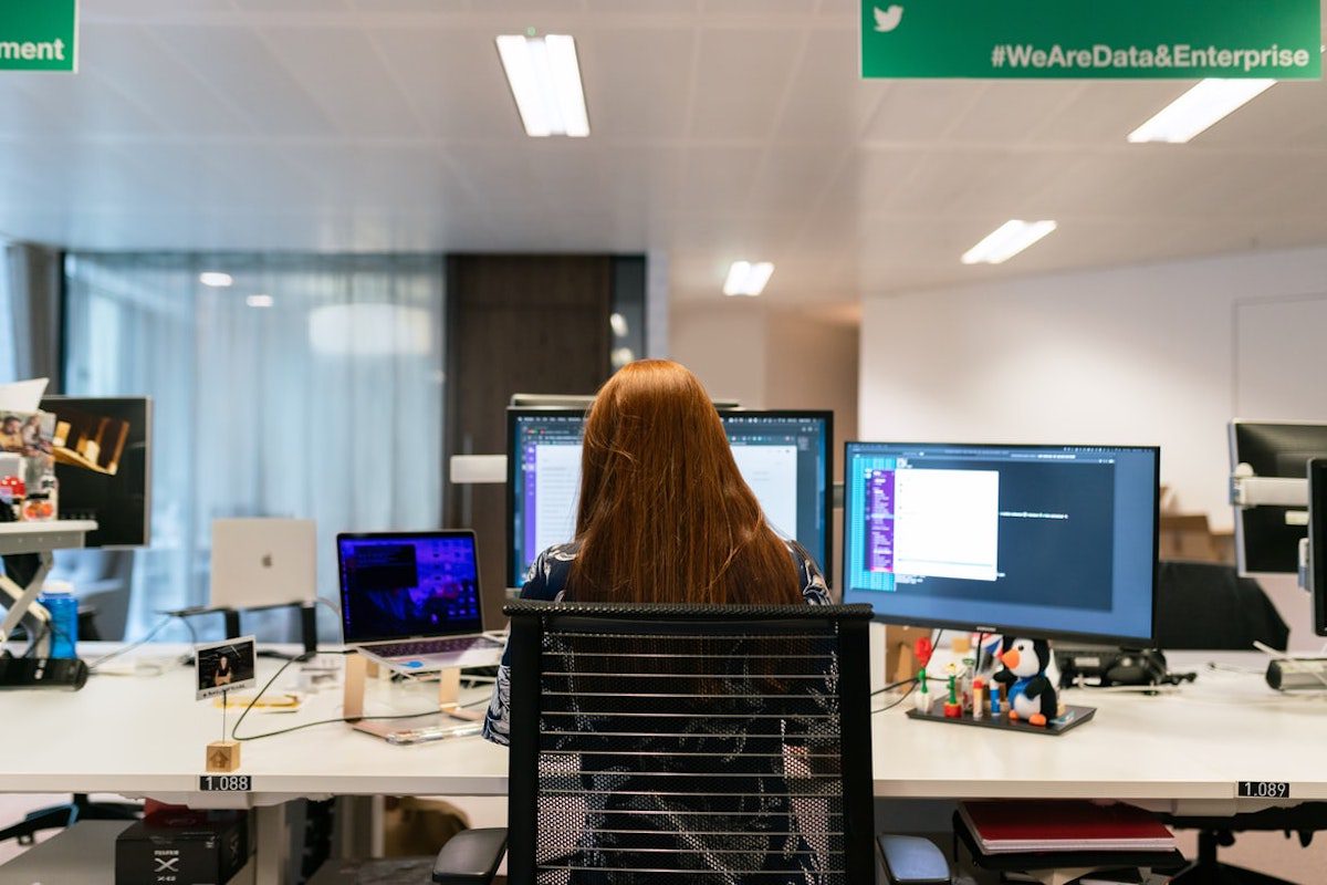 A person wearing a black printed shirt sitting in an office and working on a white desk with two computers, one laptop, and a stuffed toy on it Jobs That Use Computers