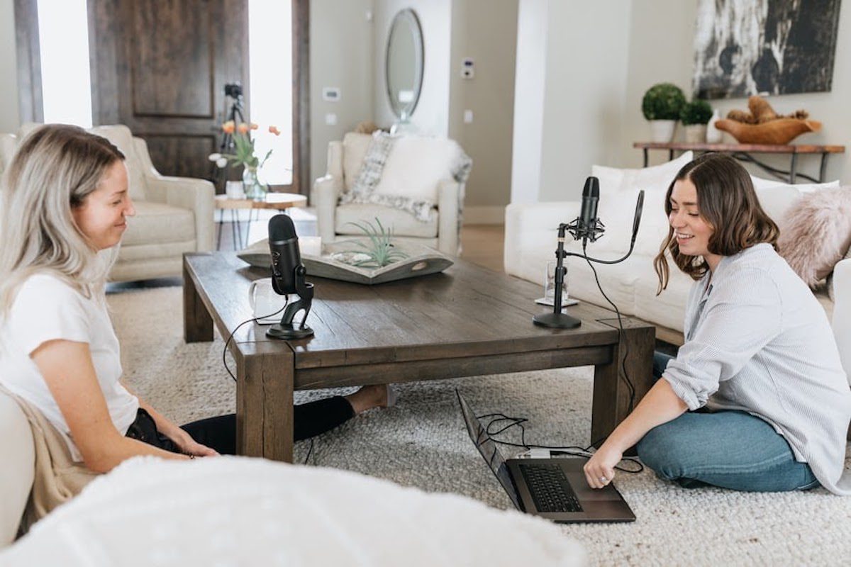 Two people wearing a white shirt and denim sitting on a living room floor and recording a podcast using a microphone and a laptop Best Podcasts For Self Improvement