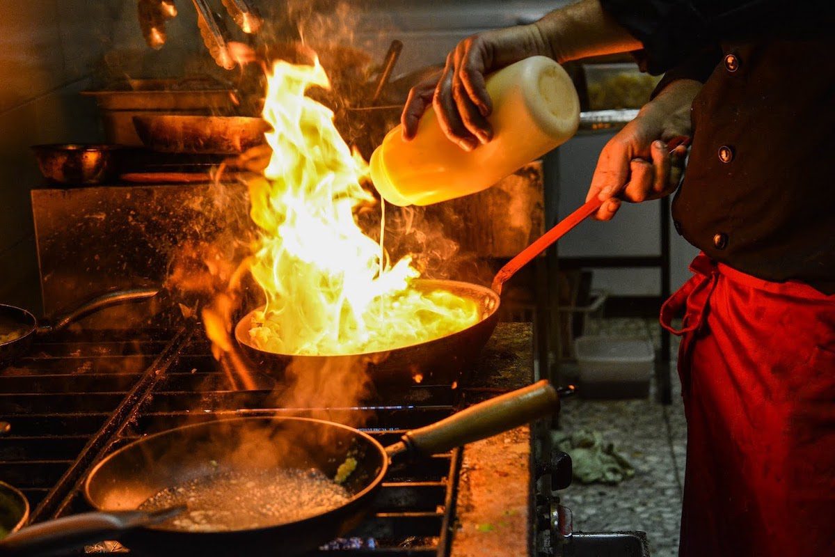 Chef wearing a red apron flambeeing seafood in a black saucepan on a gas stove.