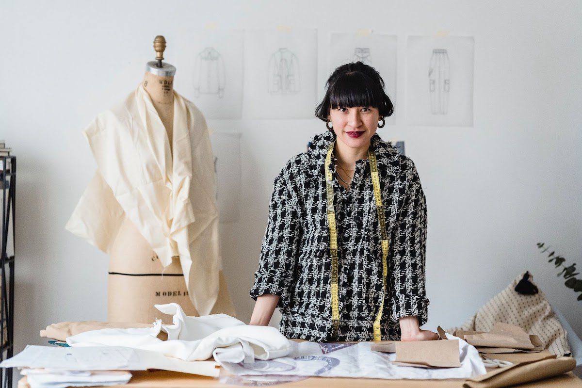 A female fashion designer standing at a table with garment sketches on a wall behind her. 