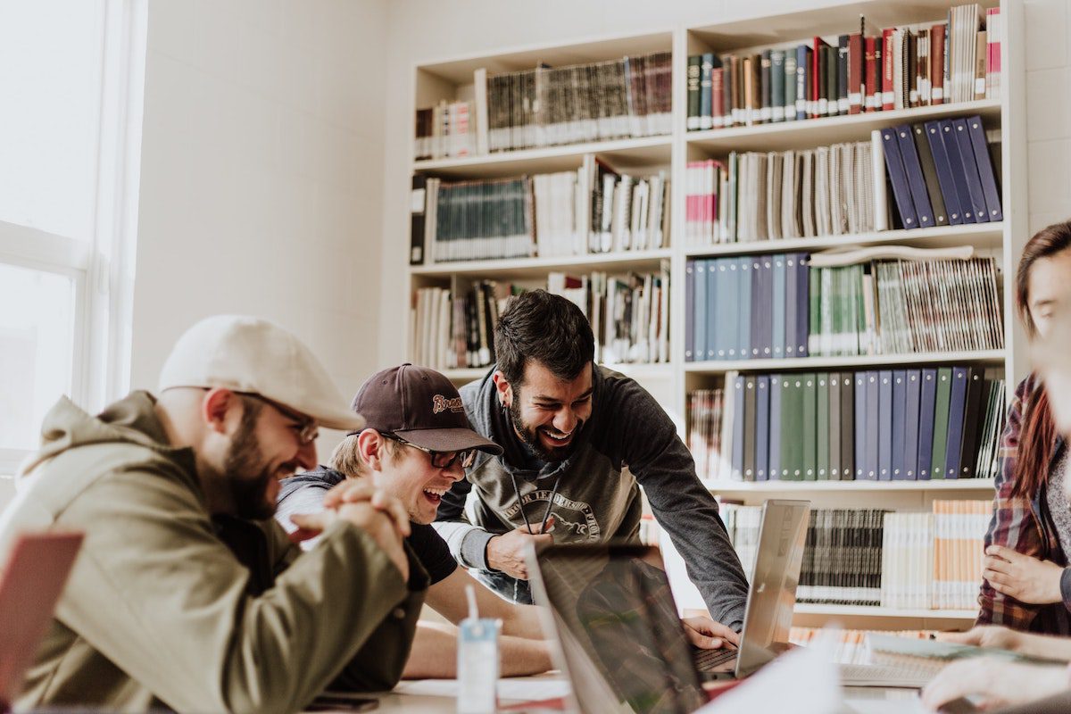 Three people laughing in a library while looking into a laptop screen. How To Get Into Graduate School