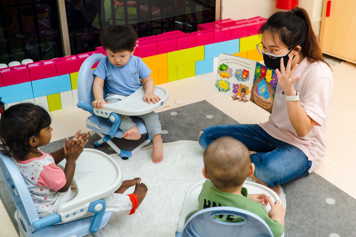 A woman wearing a facemask and glasses showing a picture book to three toddlers.